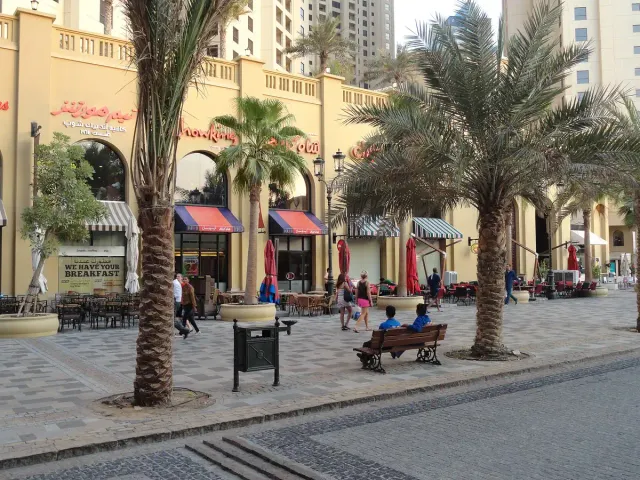 טיילת רחבה מרוצפת עם דקלים גבוהים, ספסלים ובתי קפה משני הצדדים