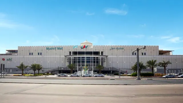 חזית ארוכה של מבנה בהיר עם דגלים, דקלים וכביש רחב בחזית