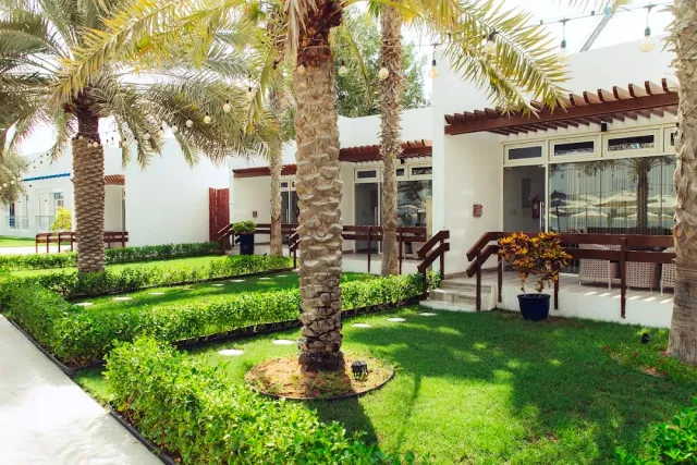 חצר מגוננת עם דקלים, שיחים מגוזמים ומבנה לבן עם מרפסת מוצלת