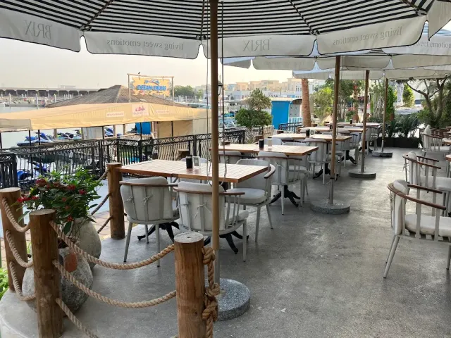 שולחנות עץ וכיסאות לבנים תחת שמשיות פסים מול מזח עם אופנועי ים