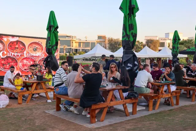 אנשים יושבים על שולחנות פיקניק בדשא עם שמשיות ירוקות ומסך גדול