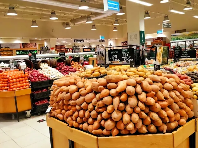 ערימת בטטות גדולה בדוכן ירקות בסופרמרקט, עם ארגזי עגבניות מאחור