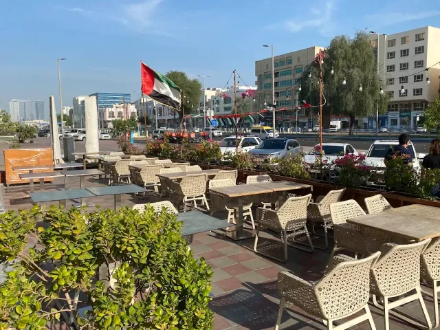 מרפסת רחוב עם כורסאות קלועות, ערוגות פרחים ודגל מתנופף מעל הכביש