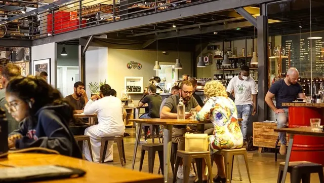 אנשים יושבים ליד שולחנות גבוהים בחלל עם גלריה עליונה ומעקה מתכת