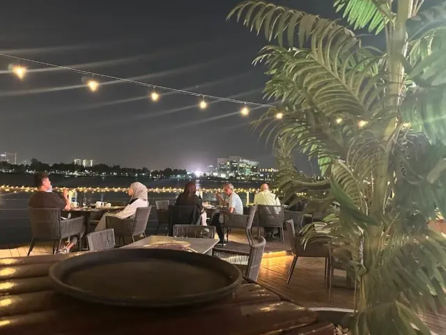 שולחנות על מרפסת עץ מול מים כהים, אורות עיר ושרשרת נורות