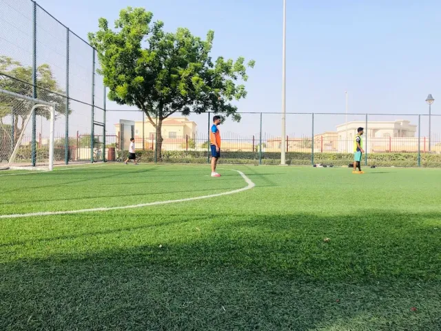 מגרש כדורגל בדשא סינתטי עם שחקנים צעירים, עץ ובתים ברקע
