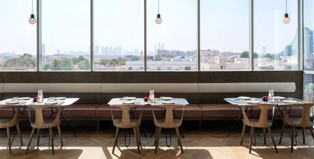 שולחנות ערוכים לאורך ספסל מרופד מול חלון גדול עם נוף לעיר